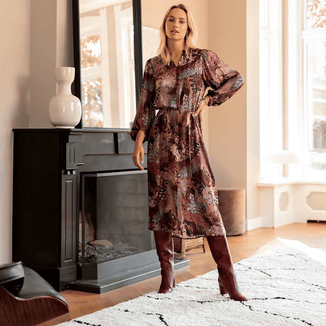 Image of a blond woman standing next to a fireplace. She is wearing a long sleeved burgundy dress and burgundy boots