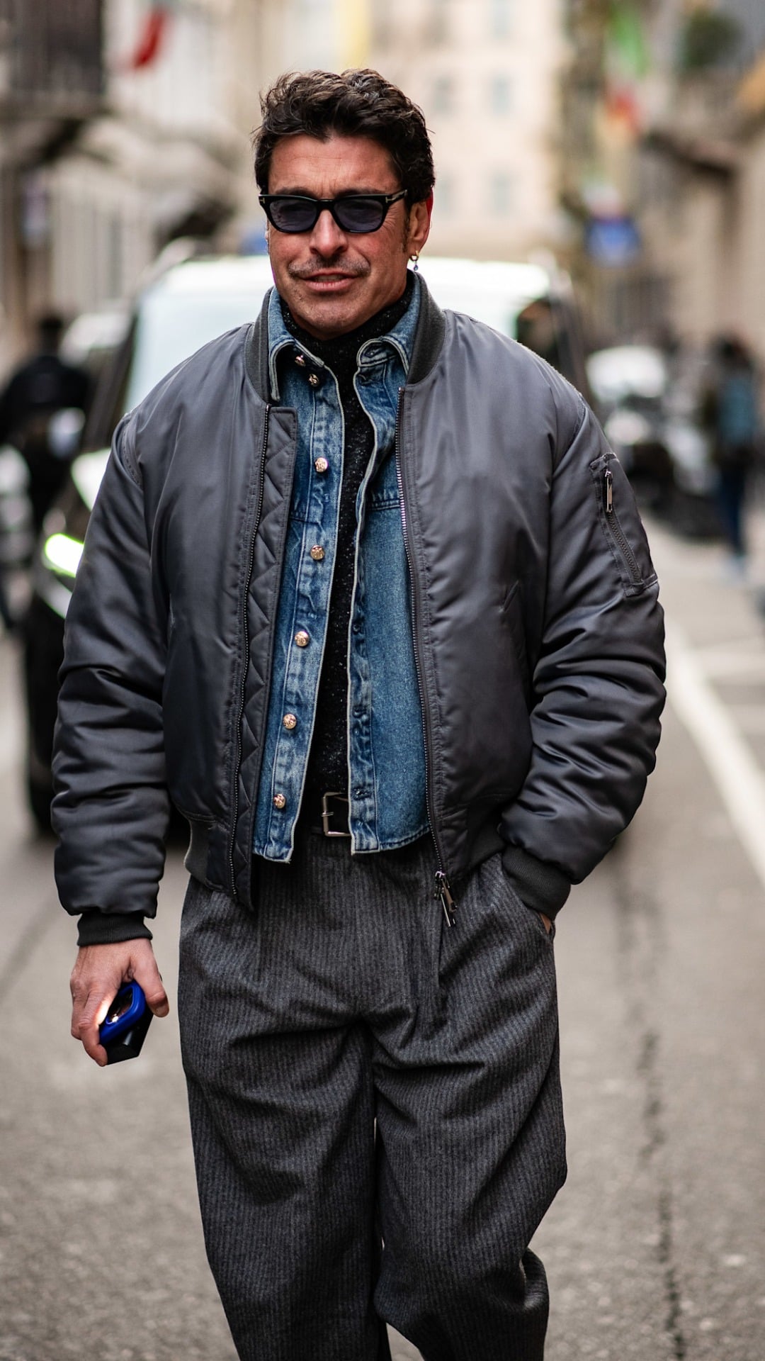 A man with an overszied grey metallic bomber jacket with a light blue wash denim jacket underneath with a roll neck base layer