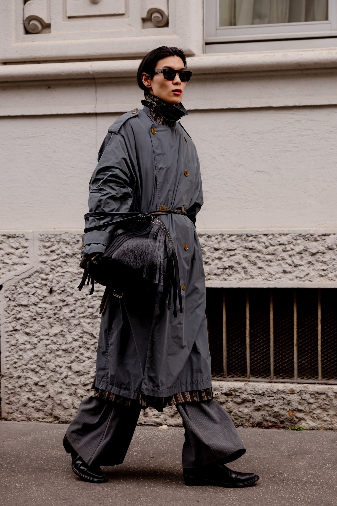 A man wearing a grey double-breasted nylon trench coat and sunglasses