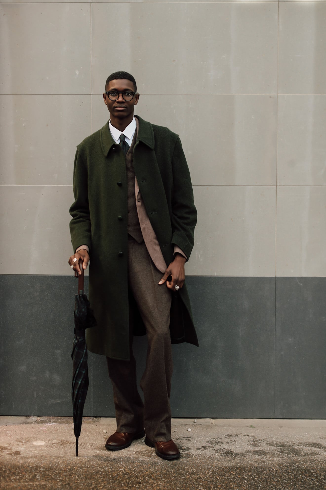 A man wearing a dark green single-breasted wool trench coat with accompanying accessories including reading glasses and an umbrella